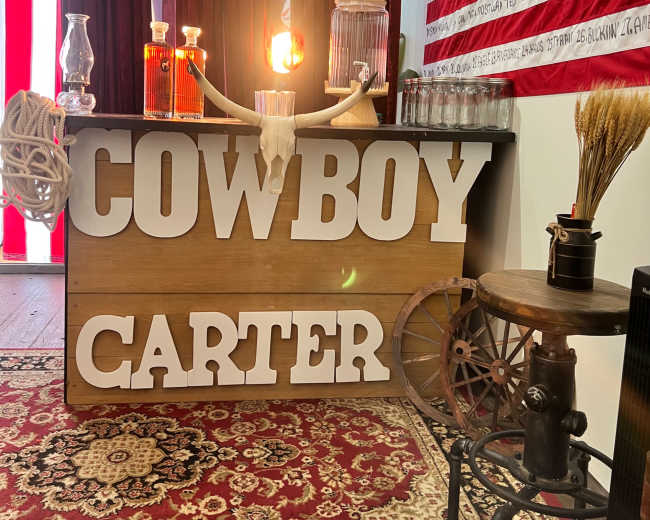 The image shows a rustic bar counter with the words "COWBOY CARTER" prominently displayed in large letters, set against a backdrop of red curtains and an American flag.