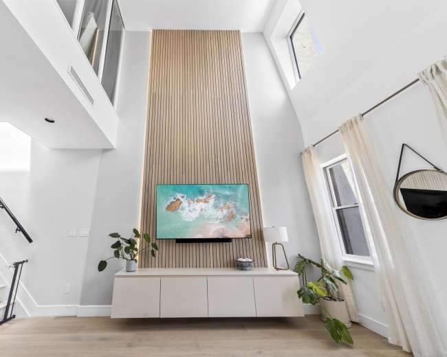 A modern living room features a high wooden accent wall with a mounted TV, light-colored furniture, and large windows.