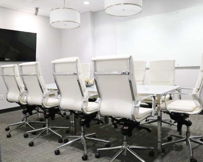 The image shows a modern conference room with a long table surrounded by white leather chairs and a large screen on one wall.