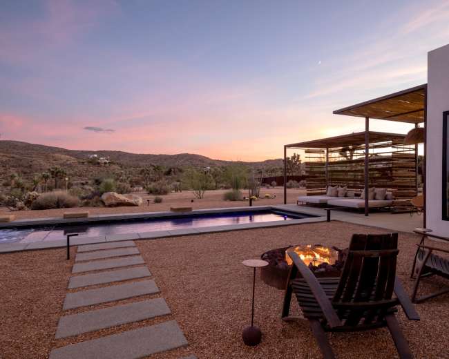 A modern outdoor space features a swimming pool, fire pit, and seating area set against a desert landscape at sunset.