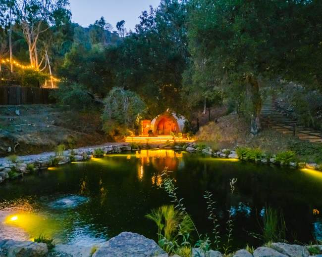 A small, illuminated structure sits beside a tranquil pond surrounded by trees and decorative lights in the evening.