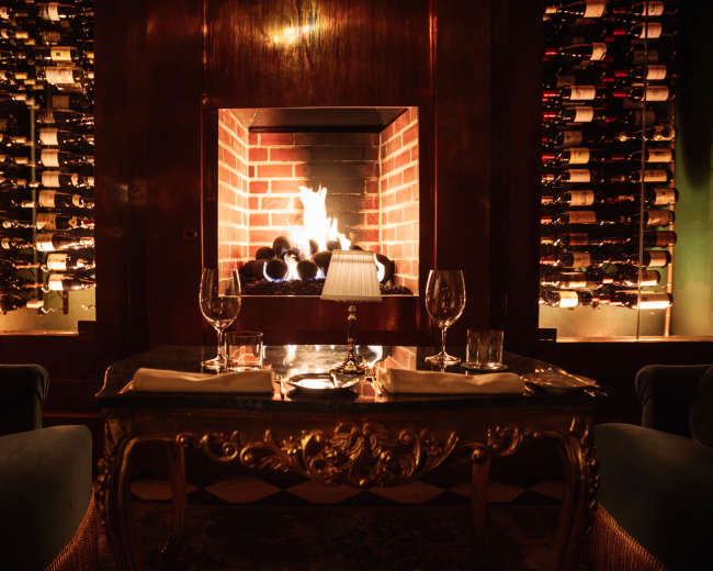A cozy restaurant setting features a brick fireplace surrounded by wine racks and a dining table with two glasses and a lamp.