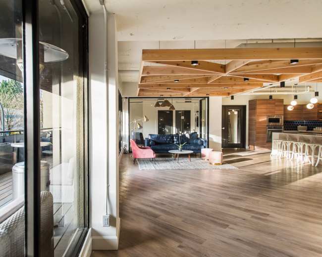 The image shows a spacious, modern interior with large windows, wooden beams, and a seating area featuring a blue couch, pink chair, and a coffee table.