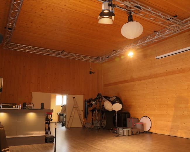 The image depicts a spacious interior studio with wooden walls, various lighting equipment, and a kitchenette area in the background.