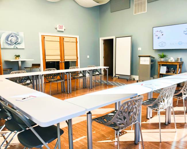 The image shows a conference room set up with tables arranged in a U-shape and chairs facing a presentation screen on the wall.