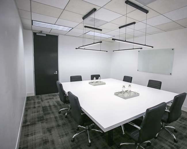A modern conference room features a large white table surrounded by black chairs, with minimalistic hanging lights and a glass board on the wall.
