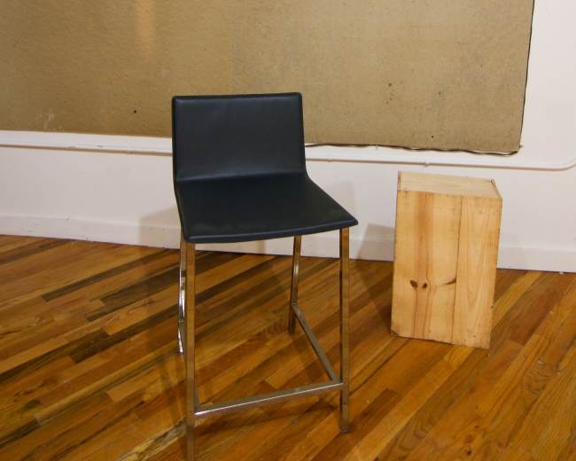 A black metal-framed chair stands in front of a large, beige textured board mounted on the wall, with a wooden block on the floor beside it.