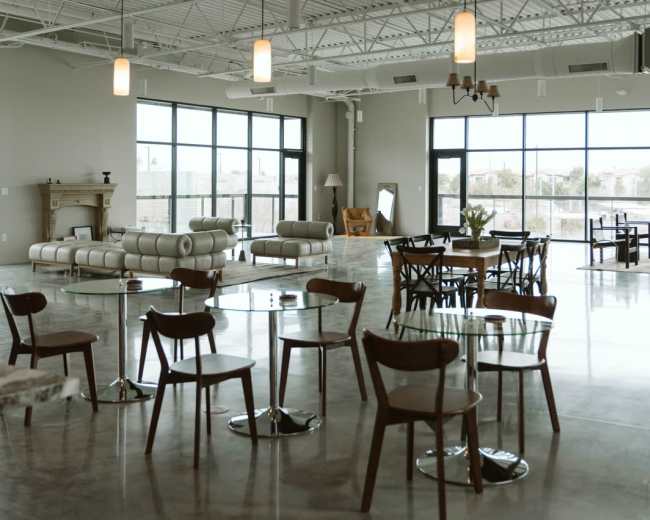 The image shows an open and airy interior space with modern furniture, large windows, and industrial-style ceilings.