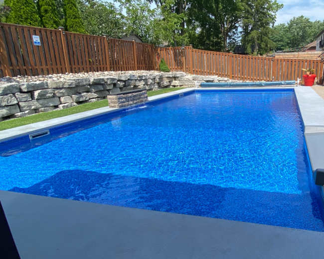 The image shows a rectangular swimming pool with clear blue water, surrounded by a stone wall and patio furniture under a shaded umbrella.