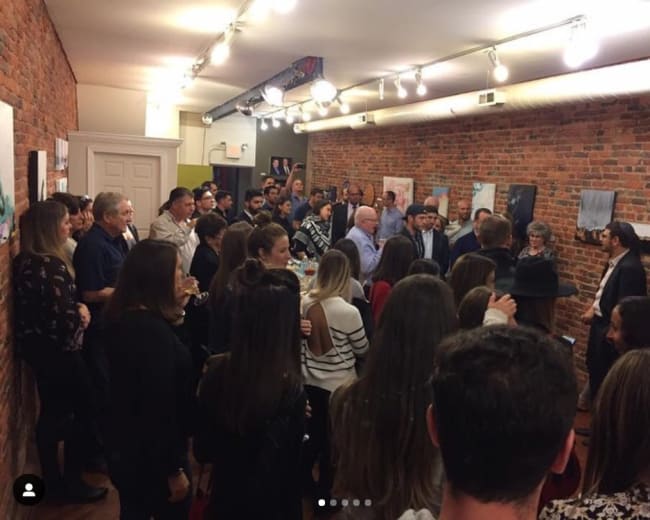 A large group of people is gathered in an art gallery with exposed brick walls, engaging in conversation while viewing various artworks.