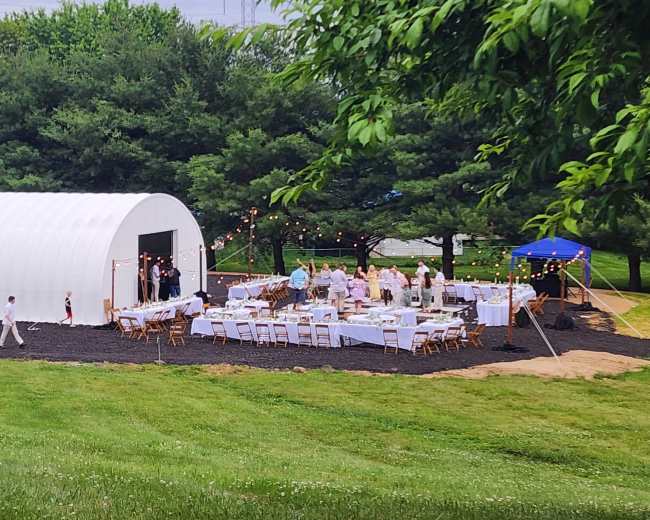 A large white barn is set up for an outdoor event, with tables and chairs arranged around a lawn area, and portable restrooms nearby.