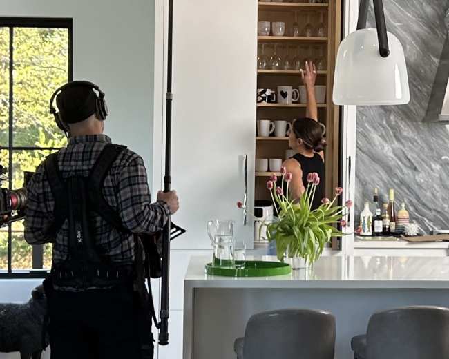 A person with a microphone on a stand records as another person reaches for glasses in a modern kitchen.