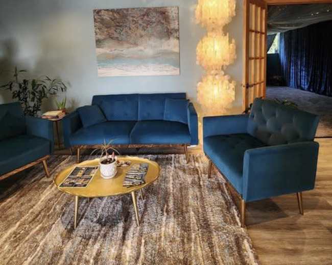 A modern seating area with teal sofas, a round coffee table, and a decorative light fixture, set against a textured rug and light blue walls.