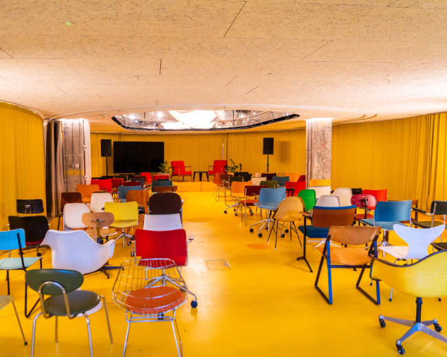 The image shows an empty circular room with colorful chairs arranged in rows on a bright yellow floor under a ceiling with indirect lighting.