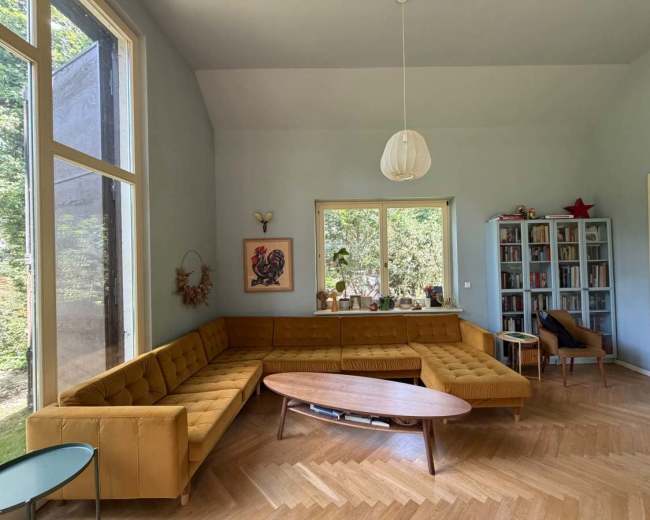 The image shows a brightly lit living room with a large mustard-colored sectional sofa, a modern wooden coffee table, and a bookshelf in the corner.