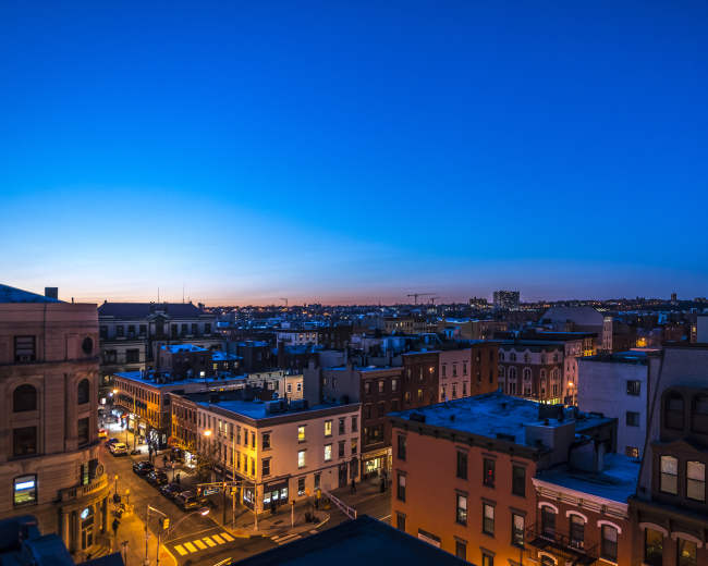 The image shows a cityscape at dusk, with buildings lining the streets and a gradient blue sky.