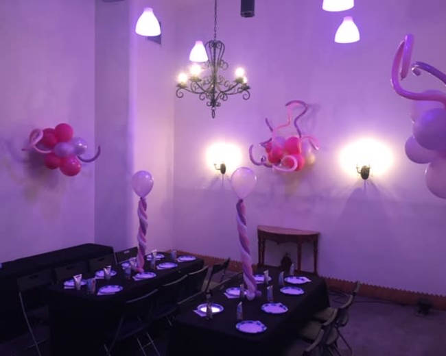 A decorated party room features a black table set for guests, complete with balloons and swirl decorations in shades of pink.