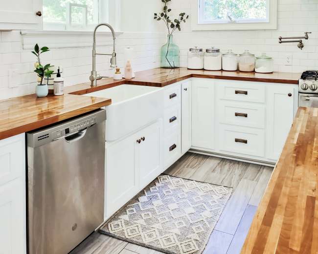 The image shows a bright kitchen with white cabinetry, a farmhouse sink, and a wooden countertop, featuring modern appliances and glass jars on the shelves.