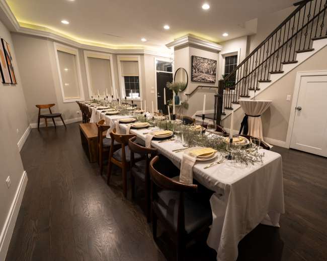 A long dining table elegantly set for a meal in a well-lit, modern room with stairs in the background.
