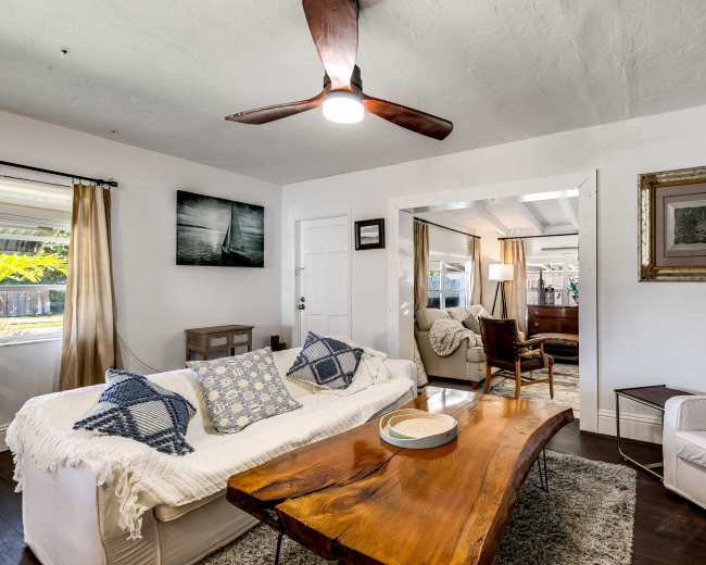 A cozy living room features a white sofa with blue patterned pillows, a wooden coffee table, and an open layout leading to another room with a desk and seating.