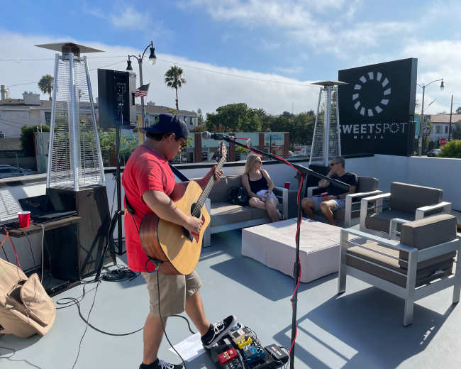 A musician plays guitar on a rooftop stage while two people sit on nearby couches.