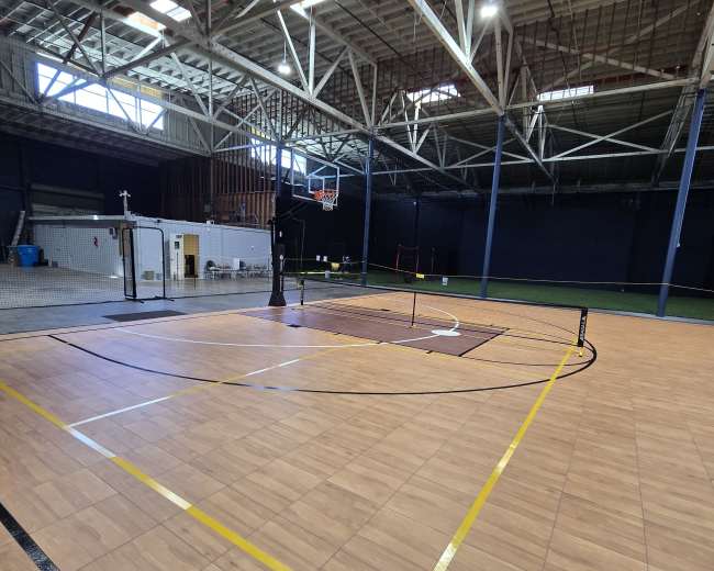 The image shows an indoor sports facility featuring a basketball court with markings for volleyball and a large open area with a high ceiling.
