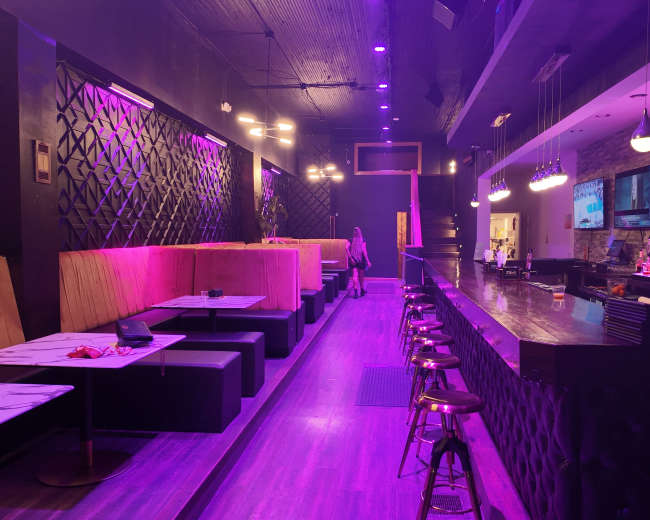 The image shows a dimly lit bar with purple lighting, featuring booth seating along one side and a long bar counter with stools on the other.