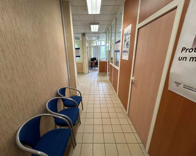 The image shows a narrow corridor with tile flooring, leading to an office at the end, accompanied by blue chairs on the left side and glass-paneled walls.
