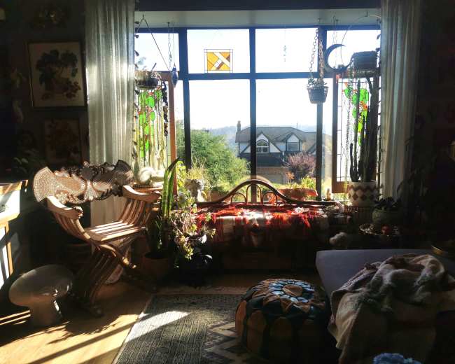 The room features a large window adorned with stained glass, offering a view of greenery and a house in the distance, while plants and decorative items are arranged around a wooden bench and a cozy seating area.