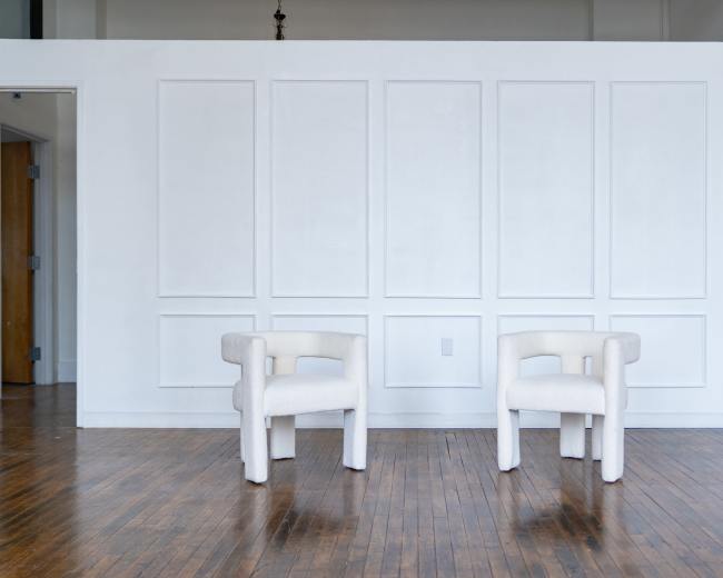 Two white, upholstered armchairs are positioned in front of a plain, white-paneled wall in a spacious room with wooden flooring.