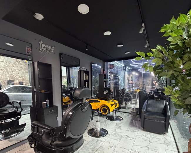 The interior of a modern barbershop features sleek black styling chairs, bright lighting, and decorative elements including potted plants and large windows.