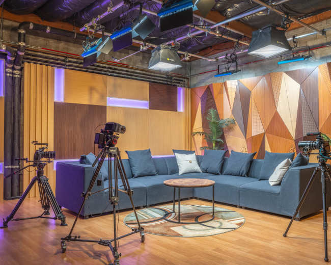 The image shows a well-lit studio setup featuring a plush sofa surrounded by multiple cameras and a round wooden table on a circular rug.