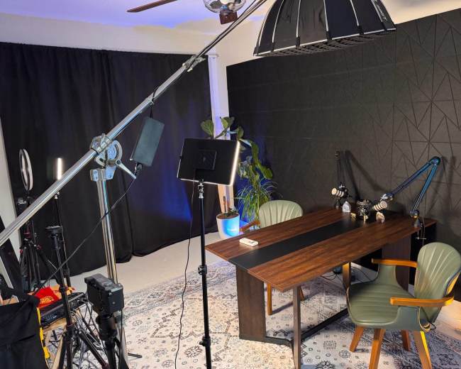 The image shows a studio setup with a desk, two chairs, lighting equipment, and a plant against a dark backdrop.