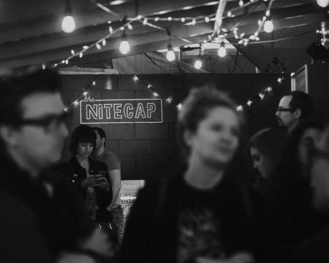 A dimly lit venue named "the Nitecap" is filled with people chatting and standing under string lights.