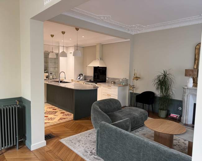 The image shows a modern living room with a green sofa, a round wooden coffee table, and a view of a kitchen area featuring white cabinets and a marble countertop.