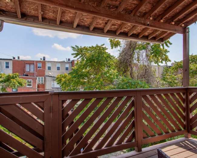 The image shows a wooden balcony with a slatted railing overlooking green trees and buildings in the background.