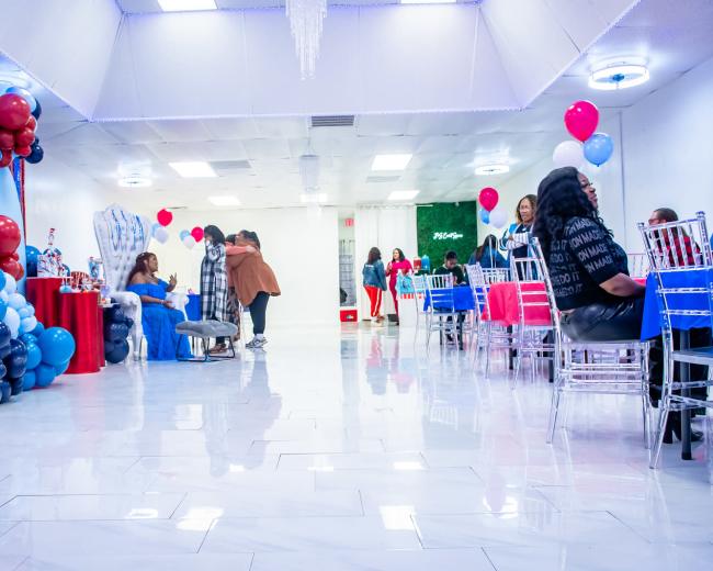 A brightly lit event space decorated with blue and red balloons, featuring tables with chairs set up for guests, some of whom are mingling and socializing.