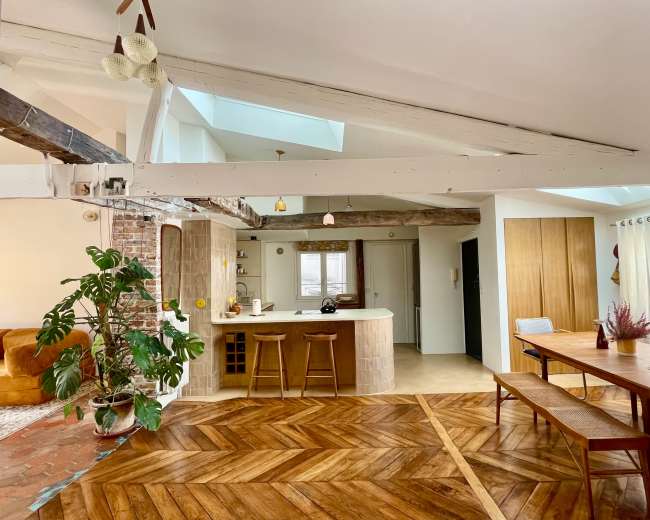 The image shows a spacious, open-concept living area with a kitchen island, dining table, and wood-paneled flooring, complemented by large windows and natural light.