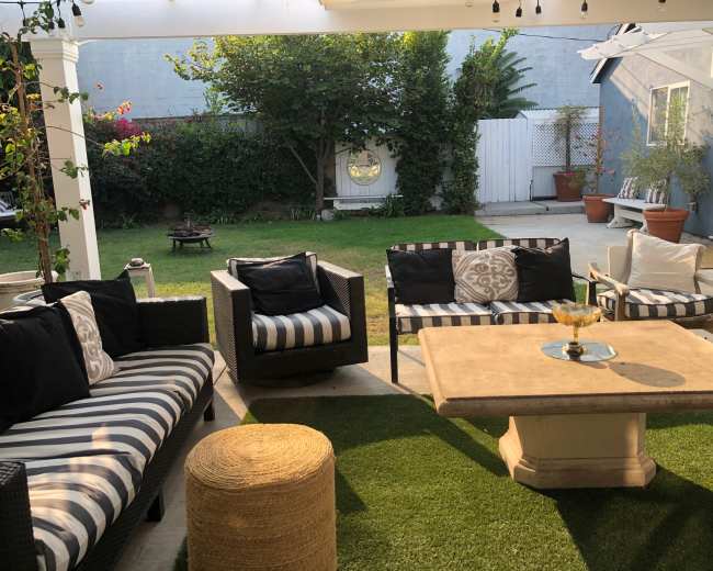 The image shows an outdoor living space with striped furniture arranged around a stone table, surrounded by greenery and potted plants.