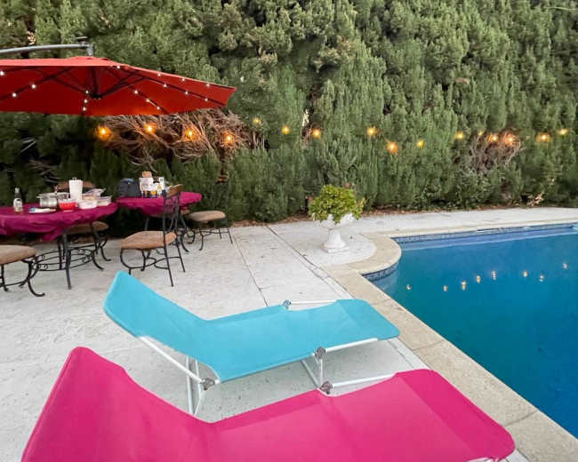 A pink and blue lounge chair are positioned beside a swimming pool, with a table set under an umbrella and greenery lining the background.