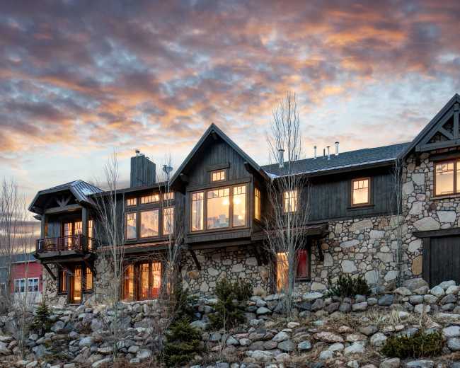 A large stone and wooden house with numerous windows sits on a rocky hillside under a dramatic sunset sky.
