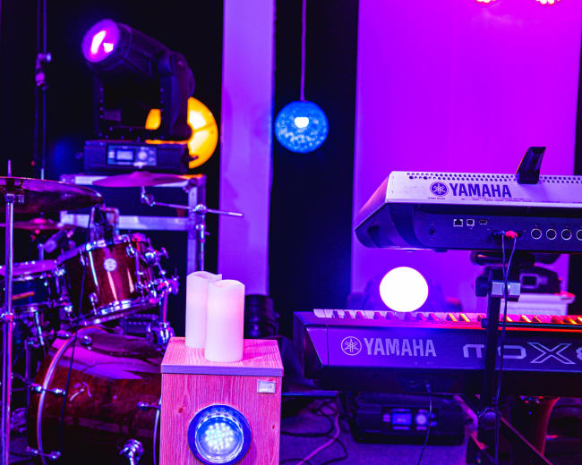A music setup featuring a drum kit, a keyboard, and various lighting elements in a well-lit studio environment.