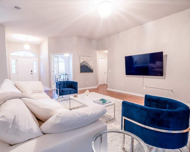 A well-lit living room featuring a white sectional sofa, two blue armchairs, a glass coffee table, and a wall-mounted television.