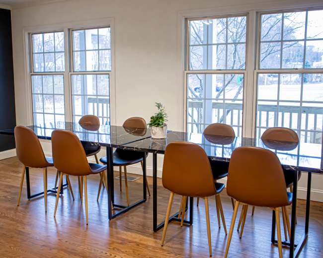 A long, dark table with eight brown chairs is set near large windows, allowing natural light to fill the room.