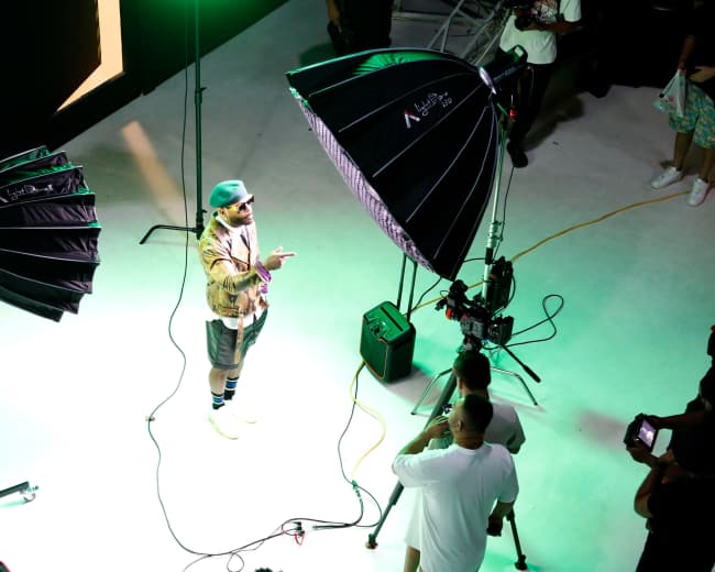 A male model stands on a lit platform surrounded by photography lighting equipment while a small crew prepares to take photos.