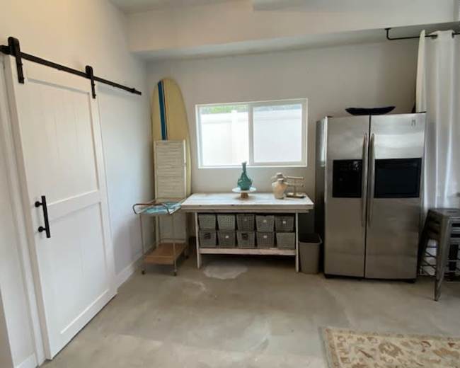A room features a sliding barn door, a stainless steel refrigerator, a countertop with storage baskets, and a surfboard leaning against the wall.