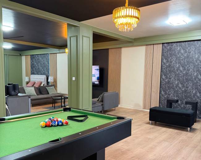 The image depicts a modern game room featuring a pool table, a seating area with a sofa, and a television against a textured accent wall.