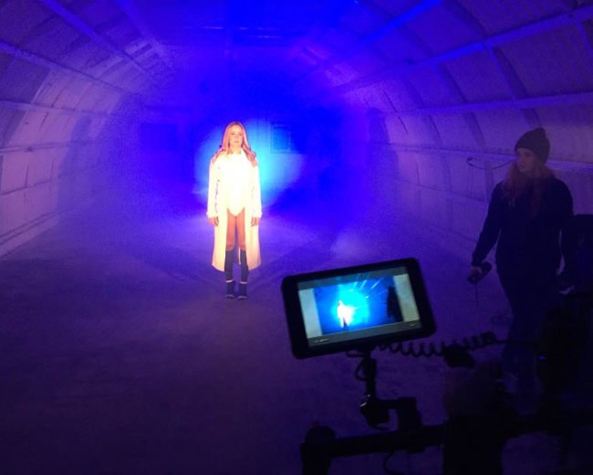 A woman stands in the center of a dimly lit, fog-filled space while a camera operator captures the scene on a monitor.