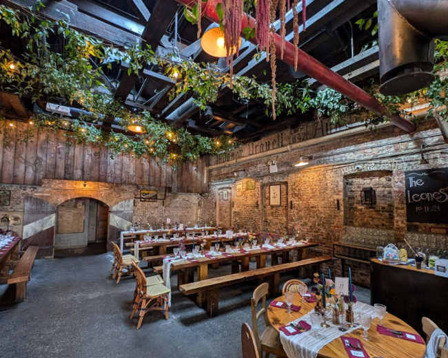 The image shows a rustic restaurant interior featuring long wooden tables set for dining, accented by hanging greenery and warm lighting.