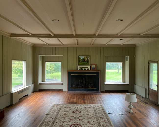 The image shows a spacious living room with wooden floors, a fireplace centered along one wall, and large windows on either side letting in natural light.